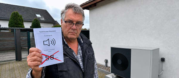 Ein Mann mit Brille steht vor einem Haus und hält ein technisches Datenblatt mit Lautsprecher-Symbol, „Außengerät“ und dem rot durchgestrichenen Wert „58 dB(A)“. Rechts daneben ist ein Außengerät an der Hauswand zu sehen.